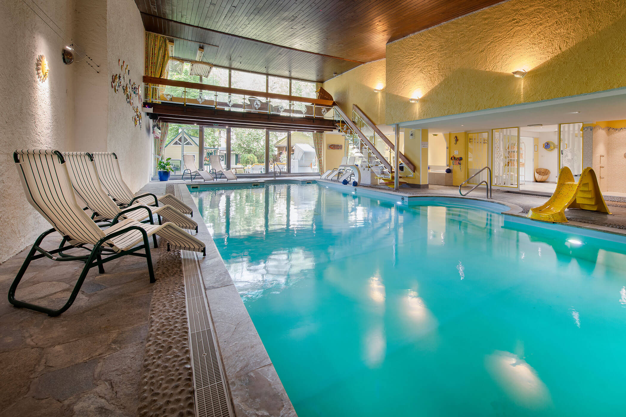 Piscina coperta in un hotel, con acqua blu e lettini a bordo, illuminata da luci soffuse e finestre panoramiche.