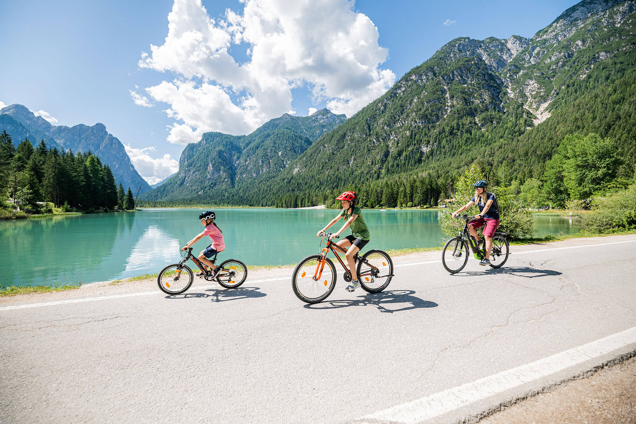 Tre persone in bicicletta su una strada vicino a un lago, godendo di una giornata soleggiata all'aperto.